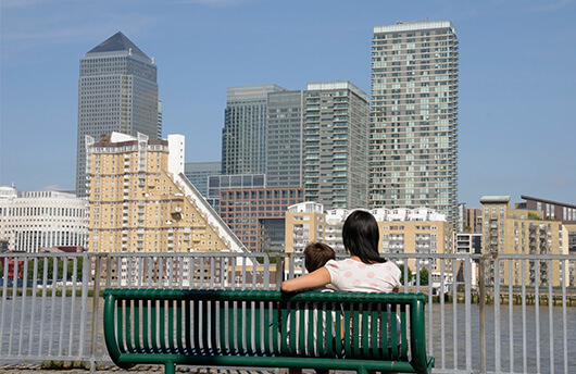 A parent and child sitting on a bench overlooking London Docklands and the River Thames
